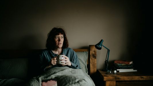 Woman sitting up in bed at night holding a mug, unable to sleep